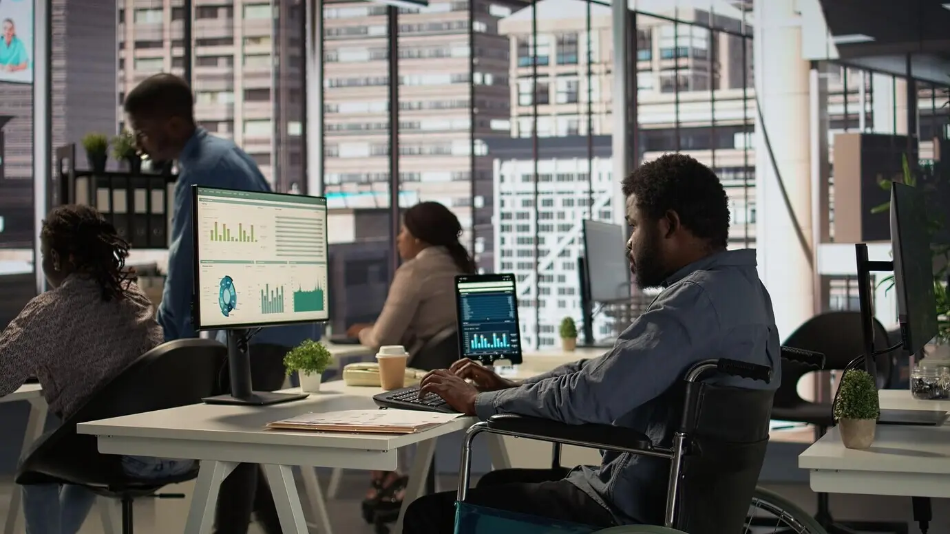 Афроамериканский сотрудник-мужчина в инвалидной коляске пытается выполнять повседневные задачи в офисе.